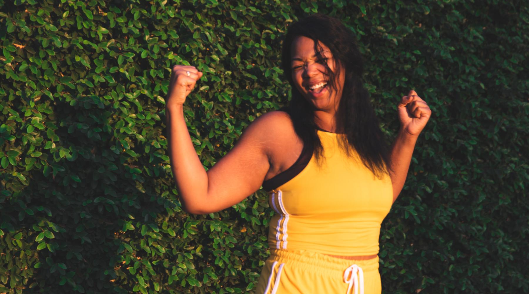 Young athletic woman in yellow tracksuit flexing biceps and smiling outdoors