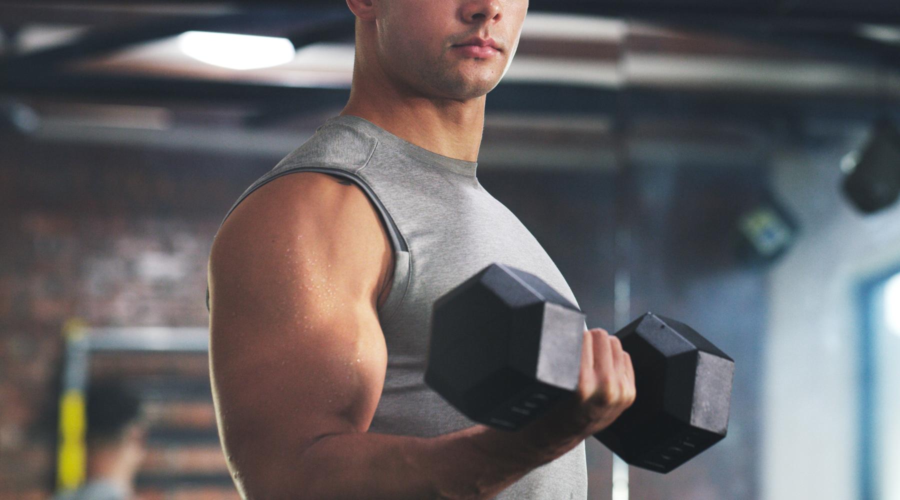 A fit man in a gray sleeveless shirt performing a bicep curl with a dumbbell in a gym, showing defined arm muscles and light sweat on his skin.