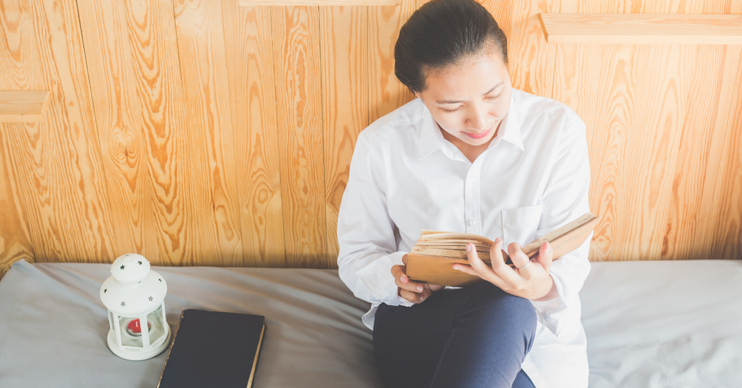 Woman sitting on bed, against headboard, reading a book.