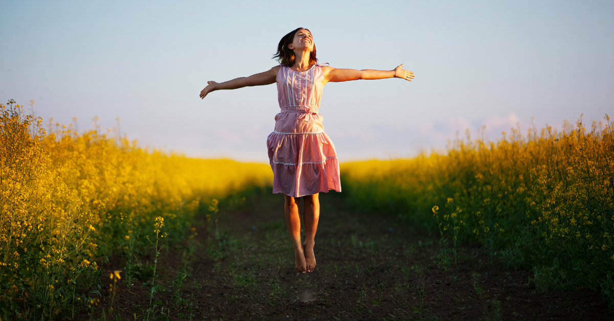 Young woman in pink dress happilly jumping in the middle of a flower field.