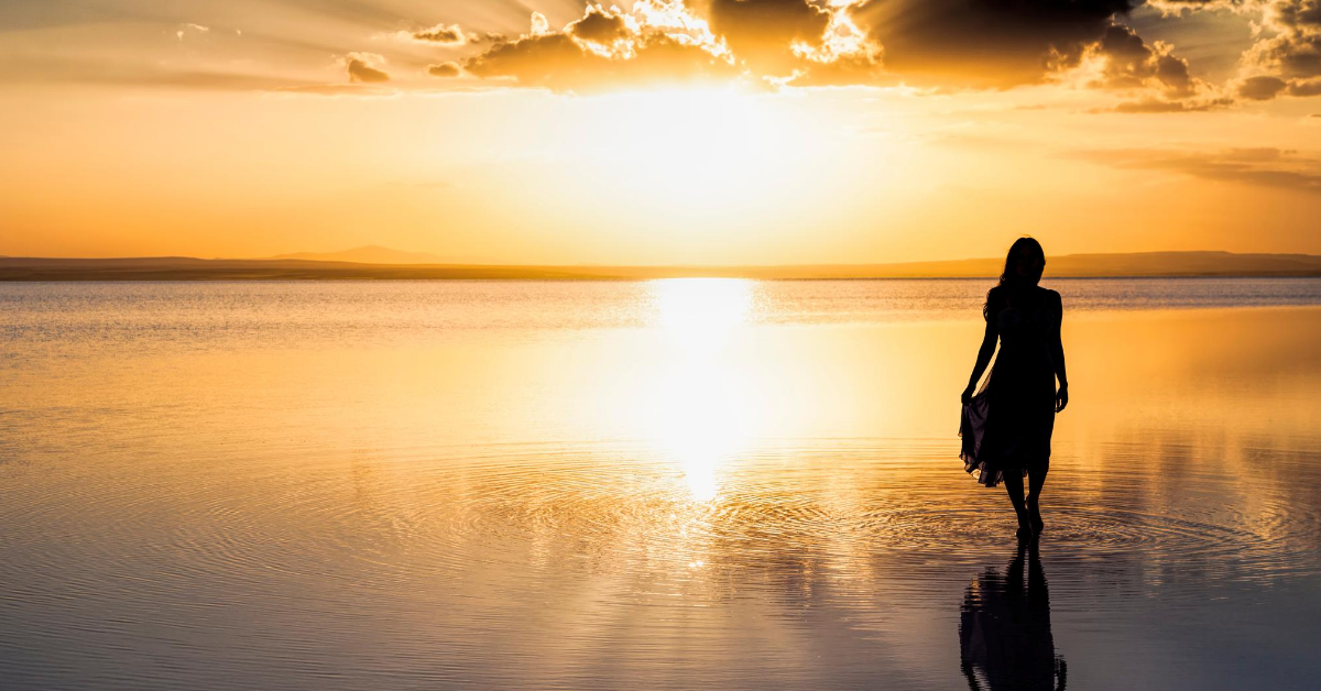 Woman walking on wet sand while sun sets in the distance.