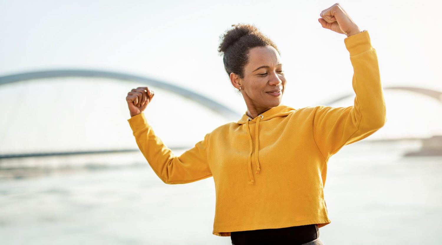 Woman in yellow hoodie flexing arm muscles outdoors