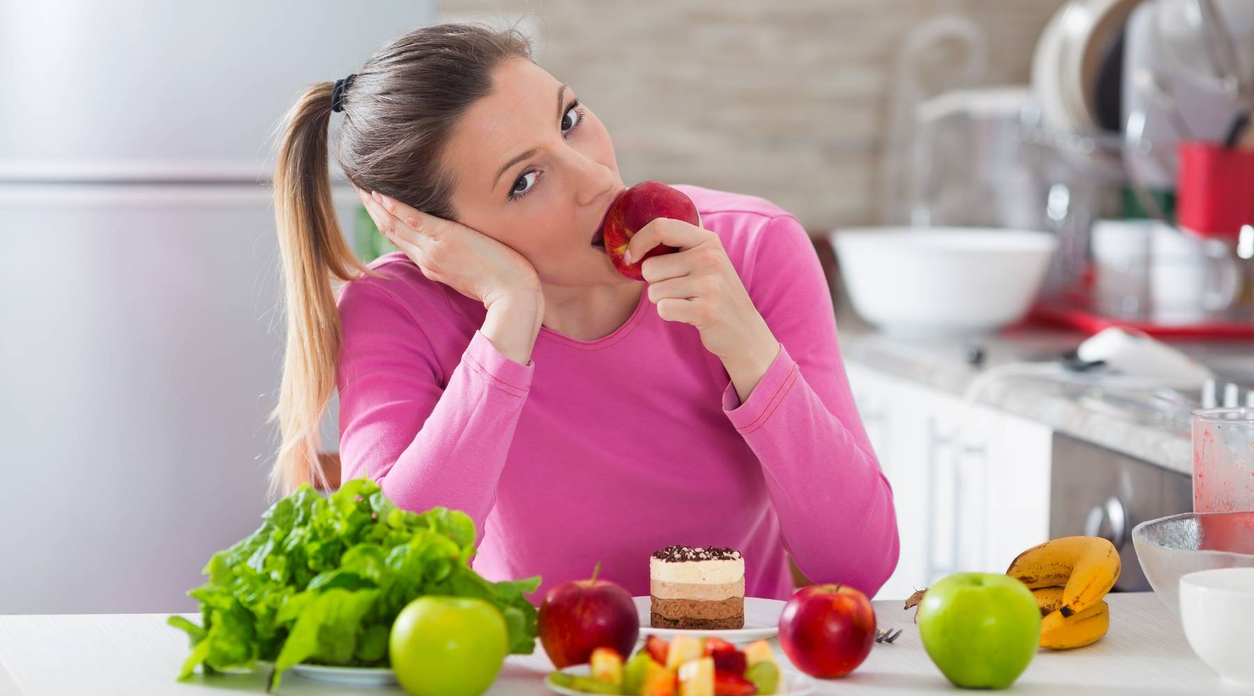 Young woman bored with her diet eating an apple while cake is standing in front of her