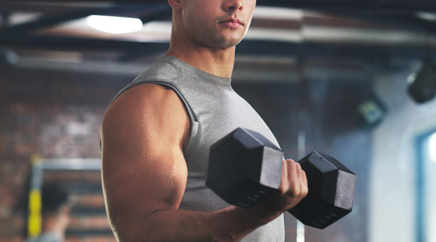 A fit man in a gray sleeveless shirt performing a bicep curl with a dumbbell in a gym, showing defined arm muscles and light sweat on his skin.