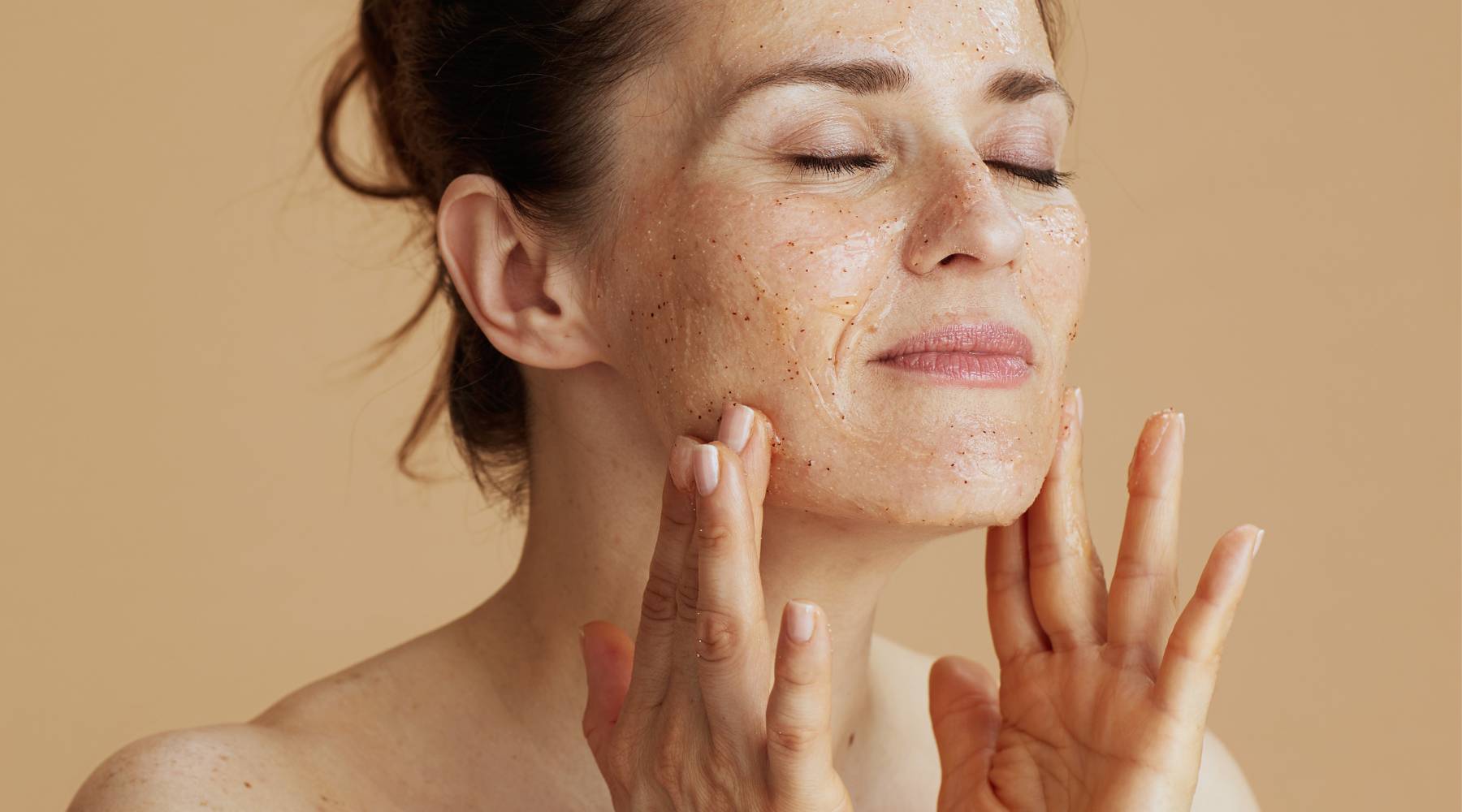 Middle aged woman applying face scrub against beige background