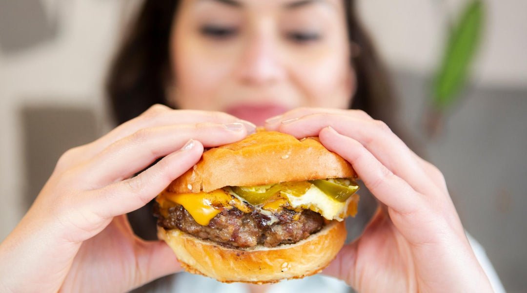 A woman holding a cheeseburger with melted cheese, jalapeños, and a sesame seed bun, about to take a bite. A blurred background suggests an indoor setting.