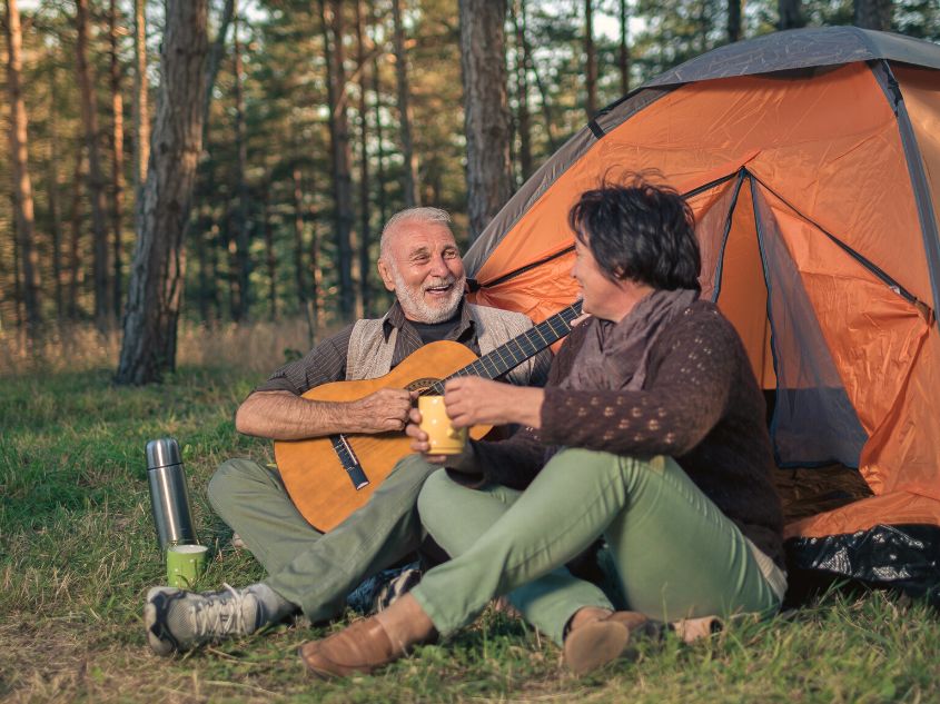 Older man and woman sitting by an orange tent outdoors. The man is smiling while playing the guitar and the woman is holding a cup of tea.