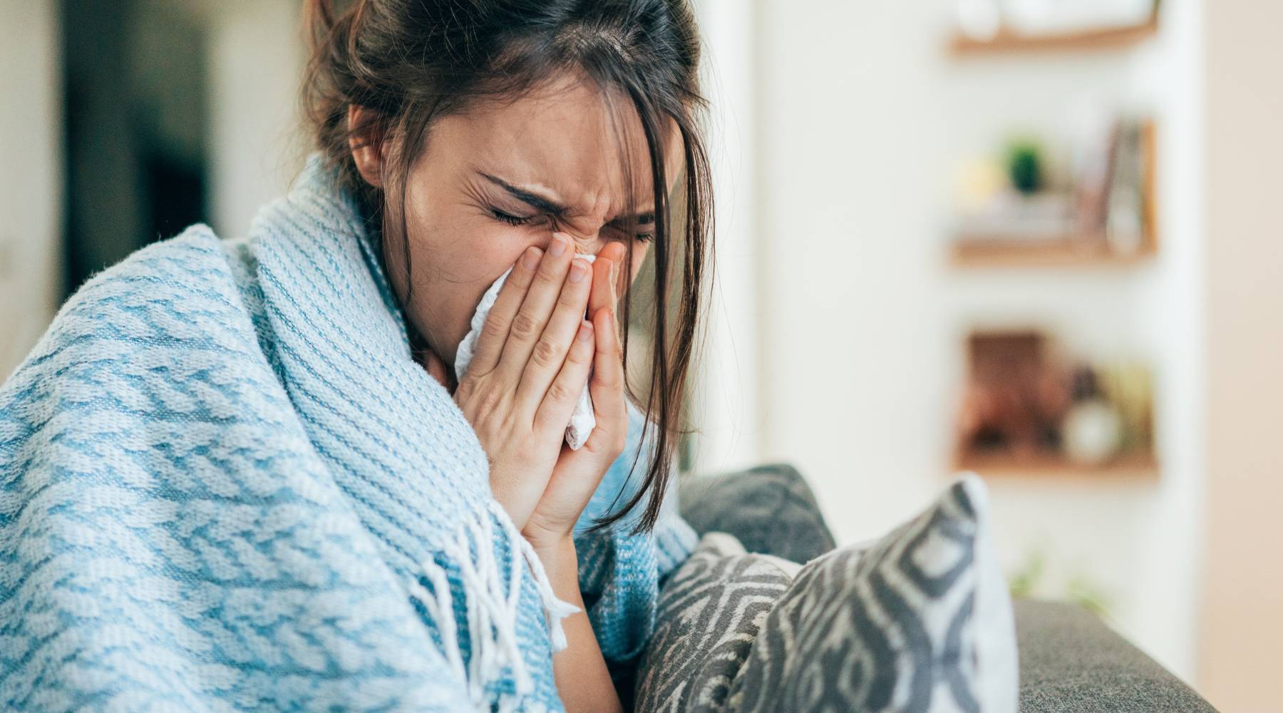 Young woman blowing her nose with a tissue while sitting covered with blanked on the sofa at home