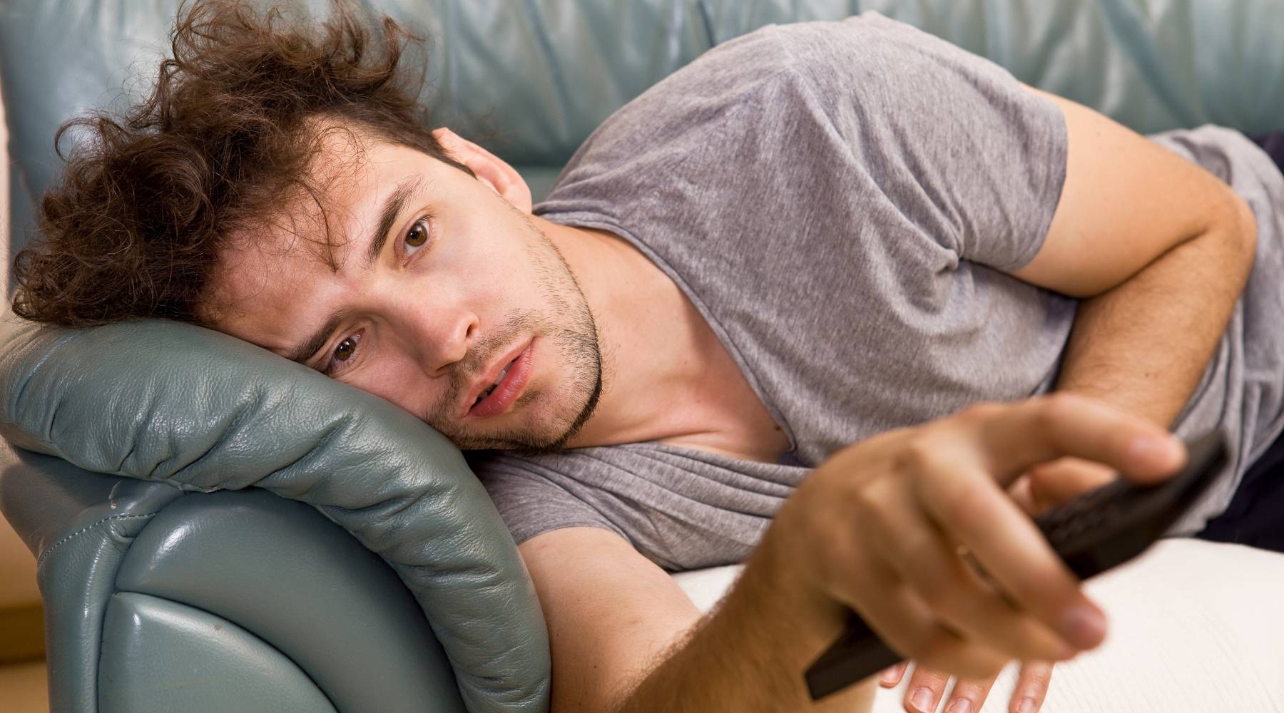 A young man lazily watches television and changes channels on the couch sedentary lifestyle health