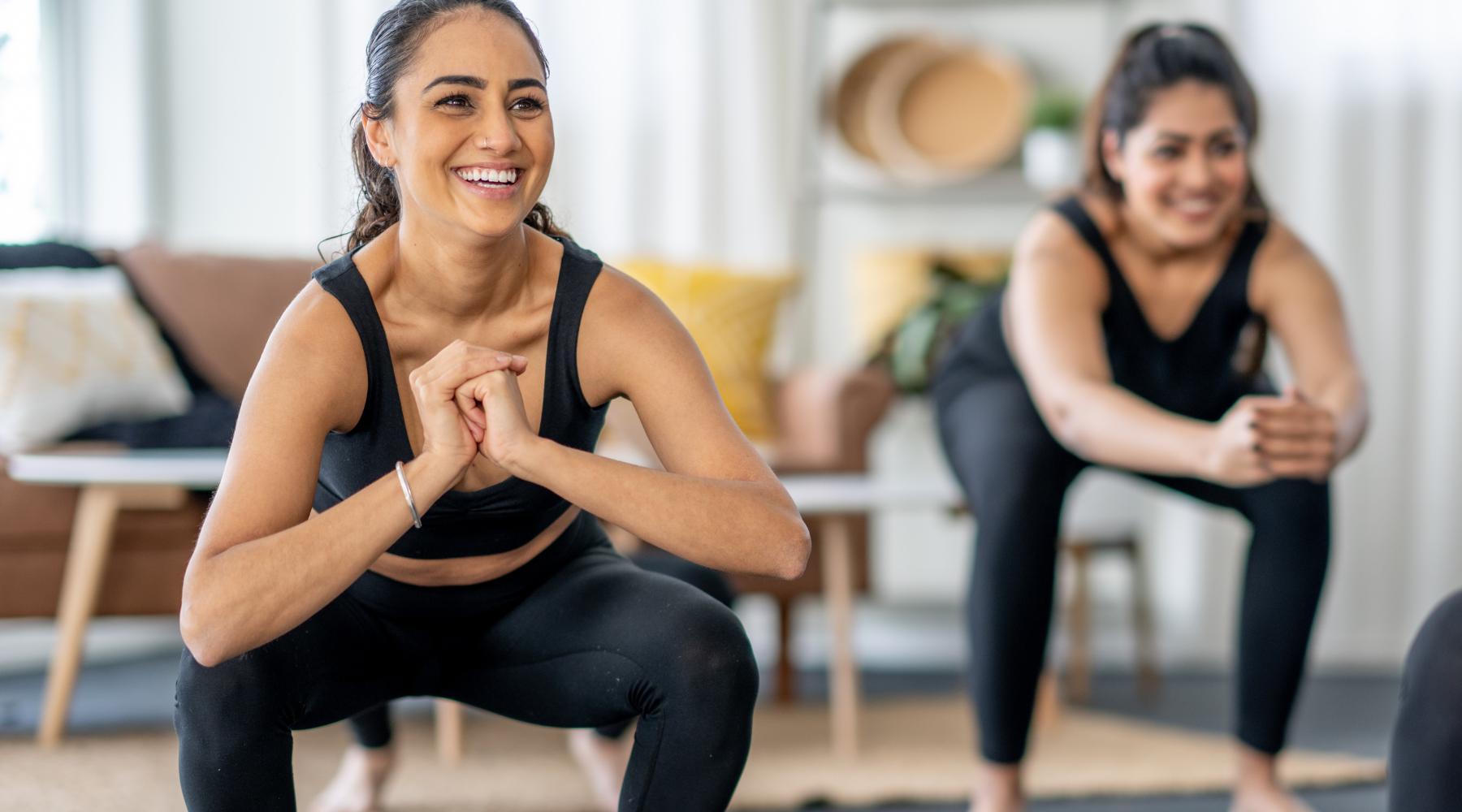 Two women in black workout outfits performing squats together in a bright living room, smiling and enjoying their fitness routine.