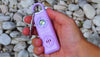 Woman's hand holding a lavender Alarmee self defense keychain. White pebbles in the background