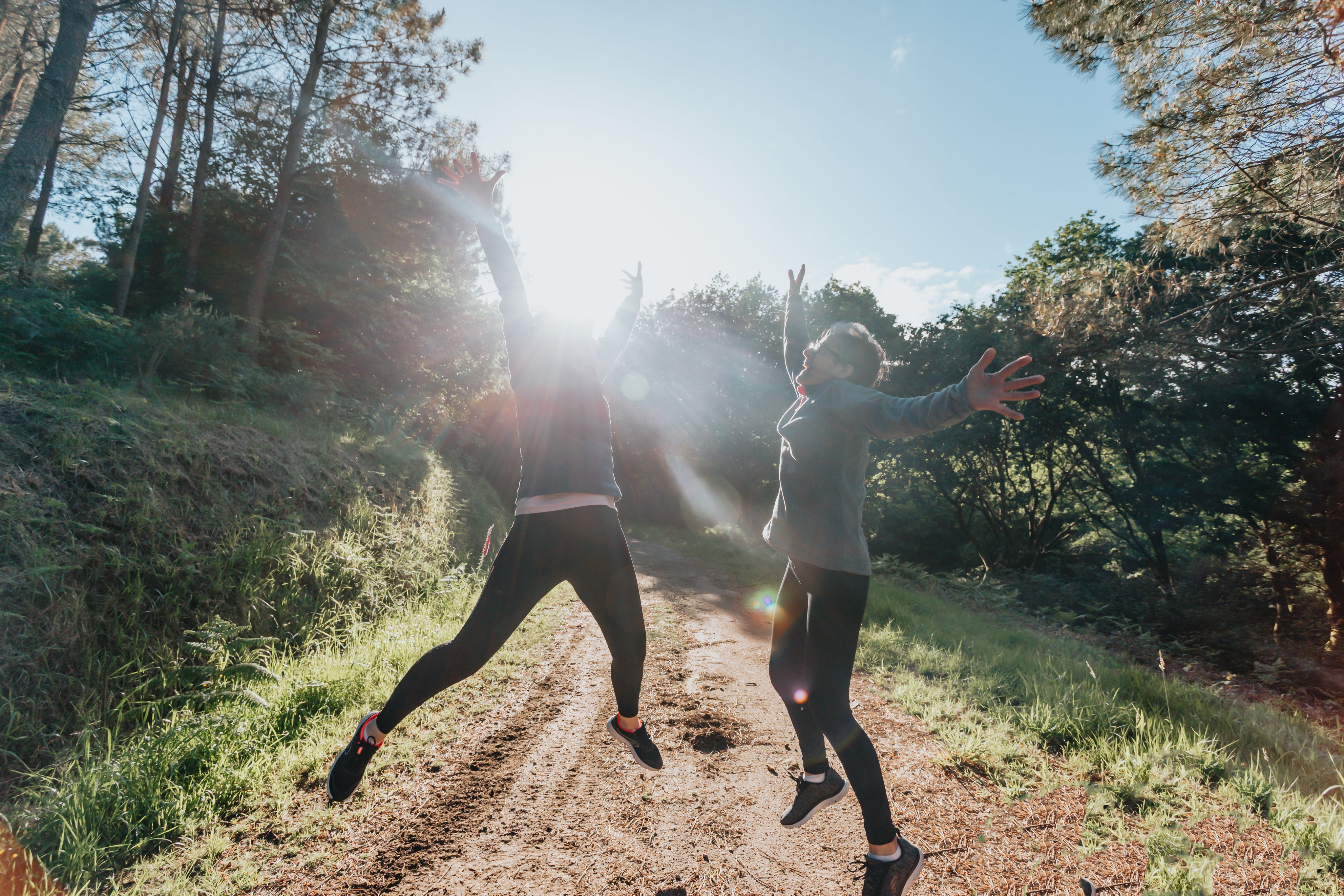 Two people in athletic clothes jumping and celebrating on a hiking trail with sunshine behind them