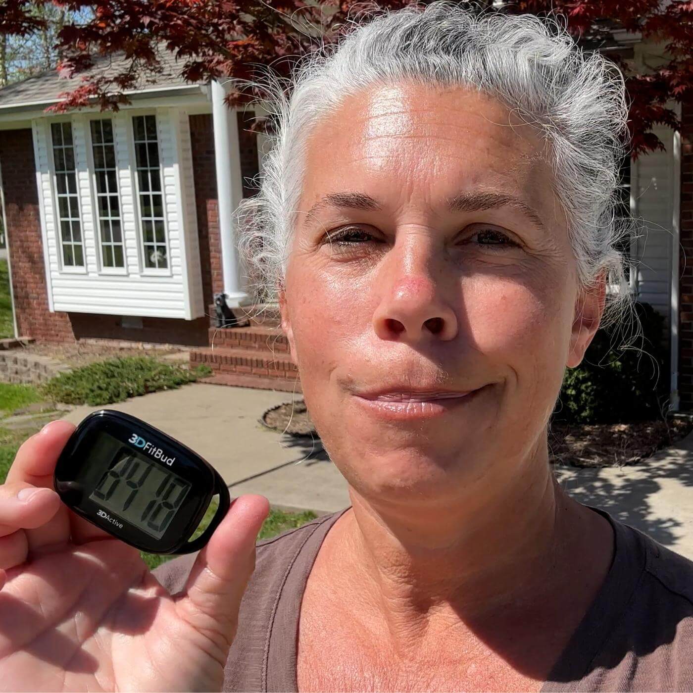 Older woman holding a black 3DFitBud Simple Step Counter outdoors in front of her house